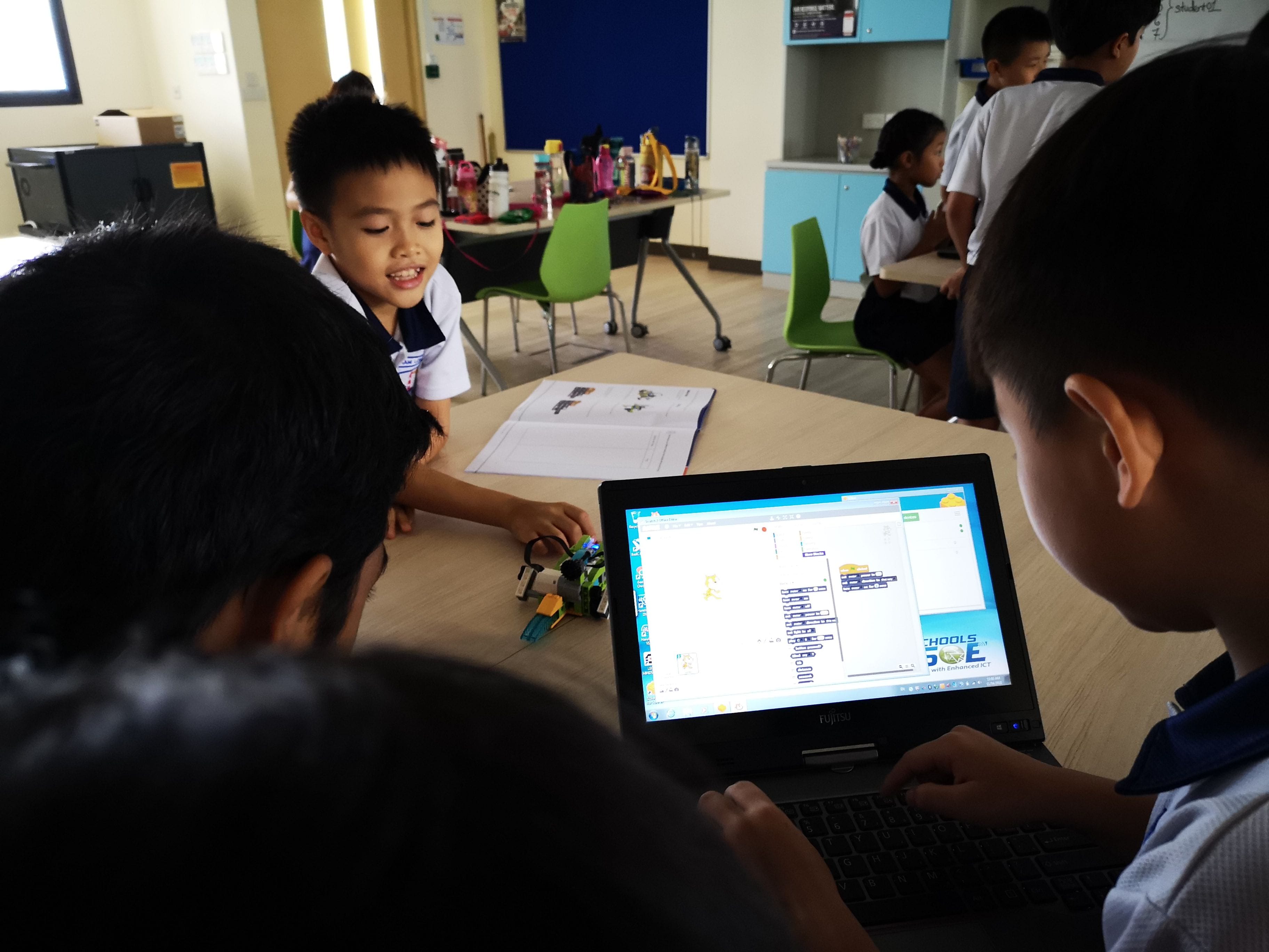 Three children in school uniforms gather around a laptop showing a block coding program, with a small robot on the desk.
